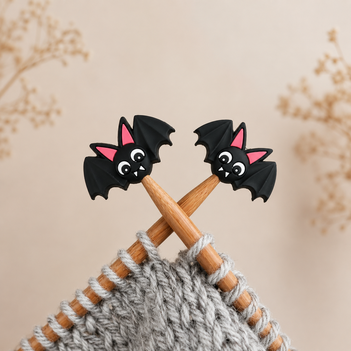 Black Bat silicone stitch stoppers with pink ears and white fangs on crossed wooden needles above grey knitting