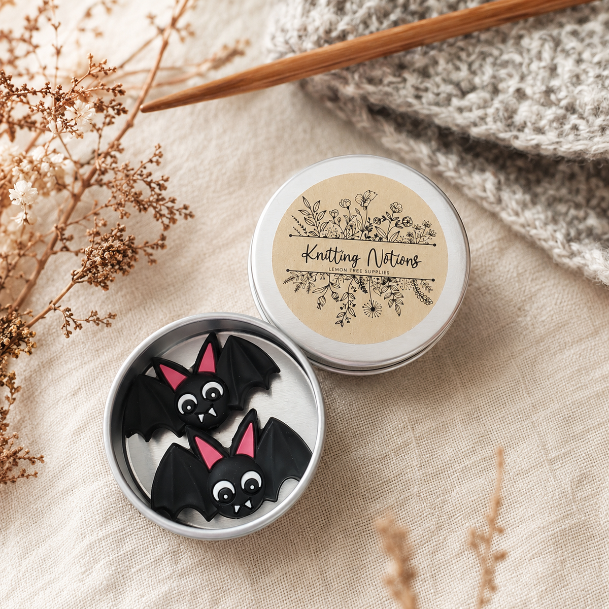 Pair of black Bat silicone stitch stoppers with pink ears in an open silver tin beside grey knitting