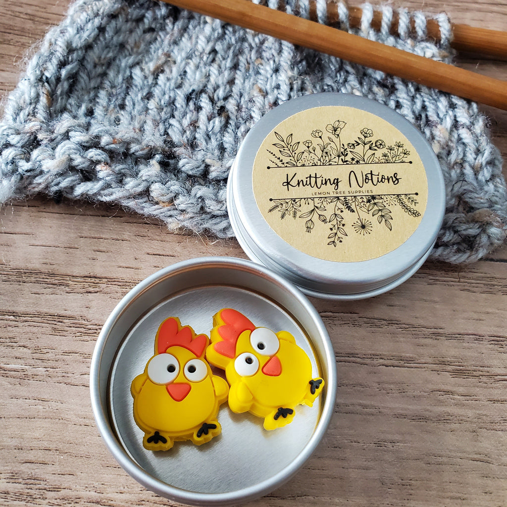 Pair of yellow Funny Chicken silicone stitch stoppers with wide eyes and red combs in an open silver tin