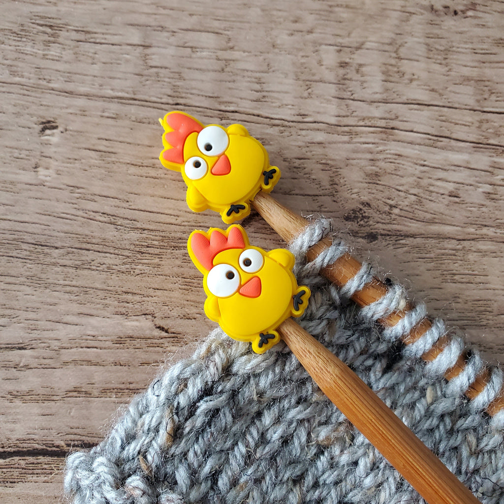 Yellow Funny Chicken silicone stitch stoppers with wide eyes and red combs on wooden needle tips above grey knitting